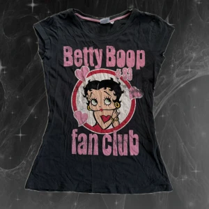 Betty Boop svart t-shirt med tryck - Svart t-shirt med stort Betty Boop-tryck och texten 'fan club' i rosa. Rund hals med rosa kant, korta ärmar och figurnära passform. Trycket har hjärtan och retrovibbar, perfekt för dig som gillar coola motiv och popkultur. S/M