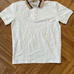 Vit Burberry t-shirt med rutig krage - Snygg vit t-shirt från Burberry med klassisk rutig krage i beige, svart och rött. Modellen har korta ärmar och knappar vid halsen. Perfekt för dig som gillar stilrena plagg med en twist från ett ikoniskt märke.