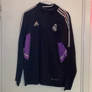 Real Madrid långärmad träningströja Adidas - Snygg svart långärmad träningströja från Adidas med Real Madrid-logga på bröstet. Tröjan har lila detaljer på sidorna, vita ränder på axlarna och halv dragkedja framtill. Tillverkad i lätt och funktionellt material, perfekt för träning eller match.