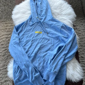 Blå hoodie ifrån Benjamin Ingrossos merch  - Säljer en chill ljusblå hoodie från Stanley/Stella i ekologisk bomull. Framtill finns broderad text 'SMILE' i gul färg som står för låtnamnet på en av Benjamin Ingrossos låtar. Hoodien har huva med snörning, magficka och långa ärmar. Perfekt för dig som gillar comfy och färgglada vibes.
