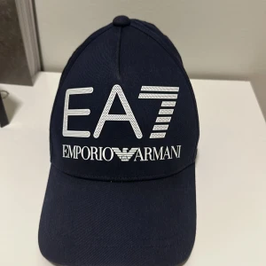 Marinblå EA7 Emporio Armani keps - Snygg marinblå keps från EA7 Emporio Armani med stor vit logga framtill och metallspänne med EA7-märke baktill. Kepsen har klassisk böjd skärm och är tillverkad i bomull. Perfekt för dig som vill ha en stilren och exklusiv look.