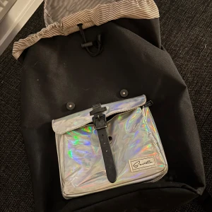 Svart ryggsäck med holografisk ficka - Cool svart ryggsäck från Santa med en stor holografisk silverficka framtill, prydd med svarta spännen och logotyp. Insidan har ett randigt foder och en praktisk innerficka. Perfekt för dig som vill sticka ut med en unik och rymlig väska.