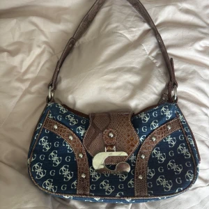   Guess jeans väska  - Snygg jeans axelväska från Guess med blått jeans  tyg och bruna detaljer i skin. Väskan har ett mönster med vita Guss-loggor, silverfärgad metallspänne och nitar. Insidan är beige med logomönster och har en praktisk inneprficka med dragkedja.