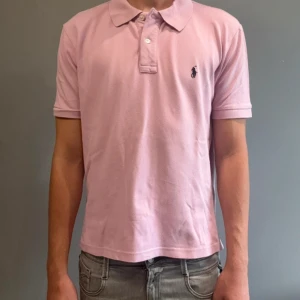 Rosa pikétröja från Ralph Lauren - Snygg rosa pikétröja från Ralph Lauren med klassisk krage och två knappar framtill. Tröjan har korta ärmar och den ikoniska broderade loggan på bröstet. Tillverkad i mjuk bomull som är skön mot huden. Perfekt för en chill och stilren look.  Storlek L men sitter som en M