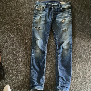 Blå slitna jeans från Diesel - Säljer ett par blå jeans från Diesel med snygga slitningar och hål på benen för en rå streetlook. Jeansen har klassisk femficksdesign, normal passform och är tillverkade i jeansmaterial. Perfekta för dig som gillar en avslappnad och trendig stil.