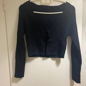 Svart croppad ribbad bolerotopp - Svart bolerotopp med croppad passform och långa ärmar. Toppen är ribbad och har en snygg v-ringning med knyt framtill. Perfekt att styla över en linne eller klänning för en trendig look. Storlek S. Aldrig använd. 