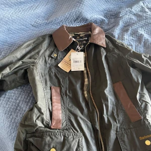 Barbour grön parkajacka med bruna detaljer - Snygg grön parkajacka från Barbour med bruna detaljer på krage och fickor. Köpte den för 4500 och har aldrig användt den, prislapp är på. Jackan har klassisk dragkedja, stora fickor med guldknappar och ett rutigt foder. Perfekt för dig som gillar tidlös stil och vill ha något som funkar hela hösten.
