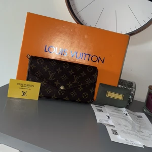 Louis Vuitton brun kuvertväska - Snygg kuvertväska från Louis Vuitton i klassiskt monogrammönster. Väskan är brun med guldiga detaljer och har insida i vinrött tyg. Kommer med matchande plånbok och kortfack. Perfekt storlek för det viktigaste och har en elegant knappstängning framtill.