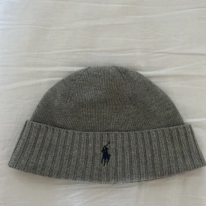  , Grå stickad mössa från Polo Ralph Lauren - Snygg grå mössa från Polo Ralph Lauren med klassisk ribbad kant och broderad marinblå logga framtill. Mössan är stickad i mjukt material och har en enkel, rundad form som passar perfekt för kalla dagar.
