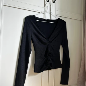 Svart ribbad topp med knyt från BikBok - Svart långärmad topp från BikBok i ribbat material. Toppen har v-ringning och snygga knytningar framtill som ger en cool och trendig look. Figurnära passform och stretchigt tyg som sitter snyggt på kroppen. 🌟