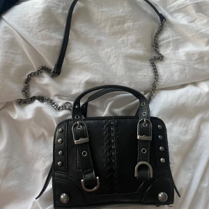 Svart handväska med kedja och nitar - Jag säljer nu min fina väska från Zara. Cool svart handväska i skinn med silverfärgade detaljer, kedja som axelrem och snygga nitar på sidorna. Väskan har två handtag, dekorativa spännen och snörningsdetalj framtill. Dragkedja upptill och rymligt fack inuti.