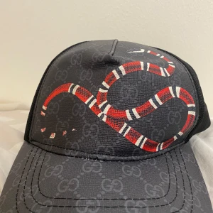 Gucci keps med röd ormprint - Snygg svart keps från Gucci med GG-monogram över hela och en cool röd och vit ormprint framtill. Kepsen har böjd skärm och justerbar passform. Perfekt för dig som vill sticka ut med en unik designeraccessoar.