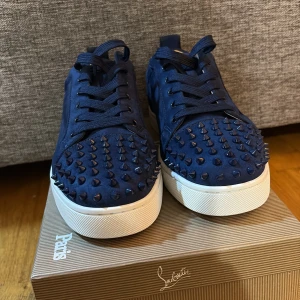 Mörkblå sneakers från Christian Louboutin - Säljer ett par mörkblå sneakers från Christian Louboutin med coola nitar över tån. Skorna har snörning, rund tå och vit platt sula. Utsidan är i mocka och insidan är ljusbeige. Perfekt för dig som gillar edgy och lyxig stil.