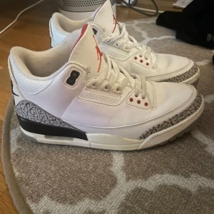 Nike Air Jordan 3 White Cement sneakers - Nike Air Jordan 3 White Cement sneakers med klassisk elefantmönster på tå och häl, vit ovandel i skinn, svarta och röda detaljer samt perforerad krage. Snygg retrostil med synlig Air-enhet i sulan och Jumpman-logga på plösen. Perfekt för dig som gillar ikoniska sneakers.