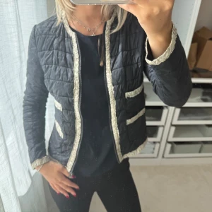 Svart quiltad jacka med beige detaljer - Quiltad svart jacka med beige kantband och detaljer vid fickorna. Jackan har rund hals, dragkedja framtill och långärmad passform. Snyggt mönstrad med raka linjer och en stilren look som funkar till många outfits. Materialet känns lätt och smidigt. Perfekt lättviktsjacka / tunn dunjacka