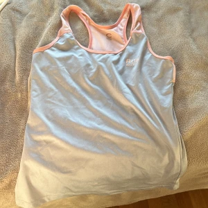 Ljusblå träningslinne Run With It - Sportigt ljusblått linne med rosa kant från Run With It. Perfekt för löpning eller gym, med racerback och mjukt syntetmaterial som andas. Snyggt tryck på bröstet och bekväm passform för aktiva dagar.