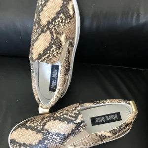 Slip-on skinn sneakers med ormmönster - Snygga slip-on sneakers från Blue on Blue med ett coolt ormmönster i beige och bruna toner. Skorna har rund tå, vit platt sula och elastiska paneler på sidorna för enkel påtagning. Perfekt för dig som gillar att sticka ut med djurmönster och vill ha bekväma skor. Väldigt dyr inköp 