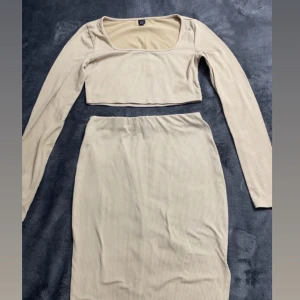 Beige ribbad croppad sett  - Snygg beige ribbad sett med croppad överdel och långärmade ärmar. Kjolen är midilång och har en elastisk midja för skön passform. Perfekt för dig som gillar stilrena och trendiga looks med en twist.