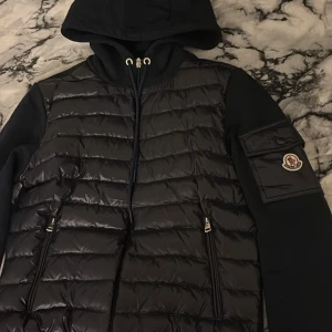 Svart pufferjacka från Moncler med huva - Säljer en svart pufferjacka från Moncler med quiltad framsida, huva med dragsko och dragkedja. Jackan har mjuka ärmar och en snygg ficka med Moncler-logga på ena ärmen. Perfekt för dig som vill ha en stilren och sportig look. Den passar båda storlk M och S
