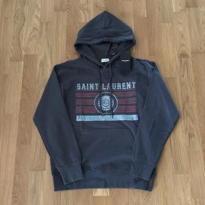 Mörkgrå Saint Laurent hoodie - Snygg mörkgrå hoodie från Saint Laurent med tryckt logga och röda ränder på bröstet. Tröjan har huva med snörning och en stor magficka. Tillverkad i mjuk bomull, perfekt för en chill och stilren look.