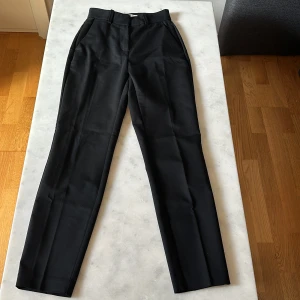 Svarta kostymbyxor från H&M, strl 32 - Svarta kostymbyxor från H&M i storlek 32. Klassisk rak passform med pressveck, bälteshällor och fickor både fram och bak. 