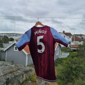 Aston Villa Mings #5 matchtröja äkta - Säljer en officiell Aston Villa fotbollströja från Kappa med Mings och nummer 5 på ryggen. Tröjan är vinröd med marinblå ränder och har ljusblå korta ärmar. Materialet är lätt och andas, perfekt för match eller träning. Snyggt AVFC-emblem och Cazoo-logga på bröstet.