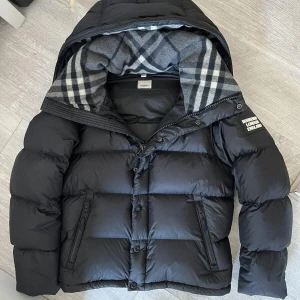 Svart dunväst/🧥 från Burberry med huva - Svart dunväst från Burberry med avtagbar huva och klassiskt rutigt foder i grått och vitt. Västen har dragkedja och knappar framtill samt två fickor med dragkedja. Märkets logga syns på bröstet. Perfekt för lager-på-lager-stil.