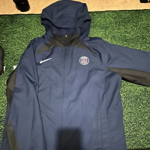 PSG Nike vindjacka marinblå  - Marinblå PSG vindjacka från Nike med huva och dragkedja. Jackan har Paris Saint-Germain-logga på bröstet och vita Nike-swoosh. Tillverkad i lätt polyester med meshfoder för extra komfort. 