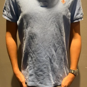 Ljusblå piké från Morris - Snygg ljusblå piké med korta ärmar och klassisk passform. Materialet är bomull med en lätt textur och på bröstet finns ett litet broderat emblem i orange. Perfekt för dig som gillar stilrena och enkla plagg med en liten detalj.