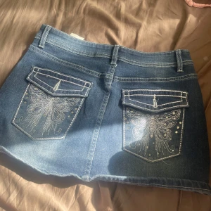 Blå jeanskjol med broderade fickor - Snygg blå jeanskjol med broderade fjärilar och silvriga detaljer på bakfickorna. Kjolen har klassiska jeansfickor fram och bak, bälteshällor och kontrastsömmar. Perfekt för dig som gillar coola detaljer och denim.