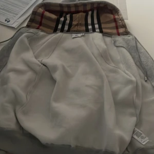 Grå hoodiejacka från Burberry M - Säljer en grå hoodiejacka från Burberry i storlek S. Jackan har klassiskt rutigt Burberry-foder i huvan och är tillverkad i mjukt bomullsmaterial. Dragkedja framtill och långa ärmar. Perfekt för dig som vill ha en stilren och lyxig streetwear-look. Passar dom som bäst till 168