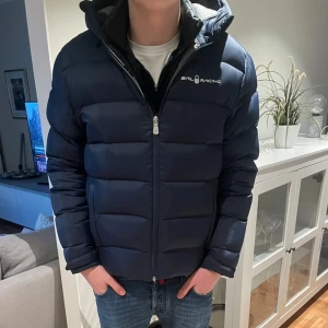 Sail Racing Jacka - Snygg mörkblå dunjacka från Sail Racing med huva och dragkedja. Jackan har en quiltad design, innerficka med dragkedja och logga på bröstet. Perfekt för kalla dagar och riktigt skön att bära. Passar dig som gillar stilren och sportig look. Fint skick utöver att hängaren lossnat men går lika bra att hänga upp den i galge eller i luvan. Går ju även att sy tillbaka den. hör av dig vid funderingar! // Ville
