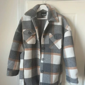 Rutig overshirt från Pieces XS - Snygg overshirt från Pieces i storlek XS med rutigt mönster i grått, vitt och beige. Jackan har stora knappar, två bröstfickor med lock och ett bälte i midjan. Perfekt för lager-på-lager och chill höst/vår-stil.