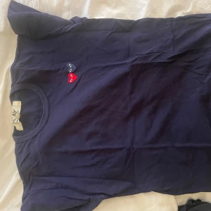 Mörkblå Comme des Garçons Play t-shirt - Snygg mörkblå t-shirt från Comme des Garçons Play med klassiska hjärtloggan i rött och blått på bröstet. T-shirten är i mjuk bomull och har en normal passform med korta ärmar. Perfekt för dig som gillar stilrena och ikoniska plagg. Skrynklig pga legat i garderoben länge! 