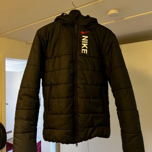 Svart Nike pufferjacka med huva M - Svart pufferjacka från Nike med huva och dragkedja framtill. Jackan har vit Nike-logga och röd swoosh på bröstet. Tillverkad i syntetmaterial med quiltad design och två sidofickor. Perfekt för kalla dagar och riktigt snygg streetstil.