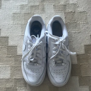 Nike Air Force 1 vita sneakers - Klassiska Nike Air Force 1 sneakers i helvitt skinn med lågt skaft. Skorna har rund tå, perforerade detaljer på tån för extra ventilation och tydlig Nike Swoosh på sidorna. Tjock, platt sula och snörning framtill. Perfekta för en clean och stilren look.