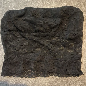 Svart spets bandeau tubtopp - Säljer en svart bandeau tubtopp i spets med blommigt mönster och vågig kant nertill. Toppen är ärmlös och har en snygg, feminin look som passar perfekt till lager-på-lager eller som statement-piece. Superstretchig och bekväm att bära.