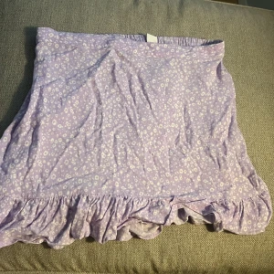 Ljuslila blommig volangkjol Lindex - Supersöt ljuslila kortkjol från Lindex med små vita blommor och volangkant nedtill. Kjolen har en mjuk resår i midjan och är gjord i ett lätt bomullstyg som känns skönt mot huden. Perfekt för dig som gillar gulliga detaljer och pastellfärger! 