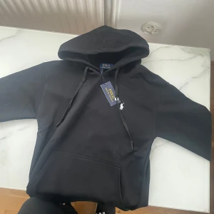 Svart hoodie från Polo Ralph Lauren - Svart hoodie från Polo Ralph Lauren med klassisk huva och snörning. Tröjan har en vit broderad logga på bröstet och en stor magficka framtill. Perfekt för dig som gillar stilrena och tidlösa plagg.