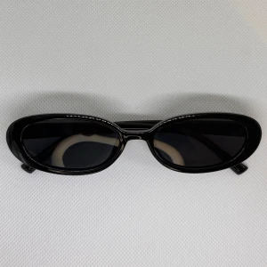 Svarta ovala solglasögon - Snygga svarta solglasögon med glansig, oval båge och mörka linser. De har en retro vibe och passar perfekt om du vill sticka ut med en stilren accessoar. Bågarna är i plast och har en smal, minimalistisk design.