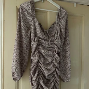 Blommig kortklänning från Nelly - Supersöt kortklänning från Nelly med lila och grönt blommönster på ljus botten. Klänningen har långärmade puffärmar, snygg snörning vid bysten och rynkade detaljer framtill och baktill som ger en cool form. Perfekt för dig som gillar romantisk och trendig stil.