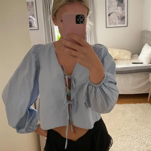 Ljusblå blus med puffärmar och knyt - Supersöt ljusblå blus med puffiga långärmar och två knyt framtill. Croppad modell som ger en chill vibe och passar perfekt till både jeans och kjol. Tunt, luftigt tyg som känns fräscht och somrigt. Perfekt för dig som gillar en feminin och trendig stil.