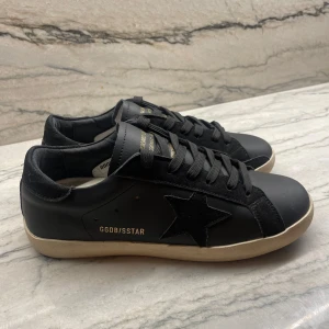 Golden Goose skor - Svarta sneakers från Golden Goose med ikonisk svart stjärna på sidan och GGDB/SSTAR-tryck i guld. Skorna har svarta snören, vit sula och detaljer i mocka och skinn. Perfekt för dig som gillar en clean men ändå snygg look.