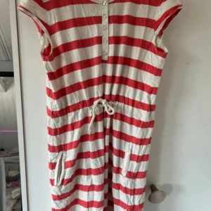 Randig rödvit kortklänning Bondelid - Säljer en kort klänning från Bondelid i storlek 160. Klänningen har breda röda och vita ränder, knappar framtill, snörning i midjan och fickor. Materialet är mjuk bomull och ärmarna är korta. Bondelid-logga broderad på ärmen för extra stilpoäng.