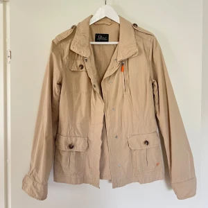 Beige vindjacka från Peak Performance - Snygg beige vindjacka från Peak Performance med klassisk krage, stora fickor med lock och knappar samt dragkedja och tryckknappar framtill. Jackan har långa ärmar med knappar vid ärmslut och coola axeldetaljer. Perfekt för vår och höst!