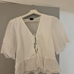 Vit blus med snörning från Gina Tricot - Superfin vit blus från Gina Tricot med v-ringning och snörning framtill. Blusen har korta ärmar och broderade spetsdetaljer längs ärmslut och nederkant. Storlek M. Nyskick! Hör av dig om du har funderingar😊
