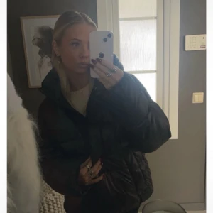 Svart pufferjacka med huva - Säljer en svart pufferjacka med huva och dragkedja framtill. Jackan har en glansig yta och breda, quiltade sektioner som ger en cool och voluminös look. Perfekt för kalla dagar när du vill vara både varm och stilren.
