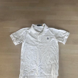 Vit Polo Ralph Lauren t-shirt slim fit - Vit t-shirt från Polo Ralph Lauren med klassisk krage och korta ärmar. Slim fit-modell med broderad marinblå logga på bröstet. Tillverkad i mjuk bomull som känns skön mot huden. Perfekt för dig som gillar stilren och enkel design.