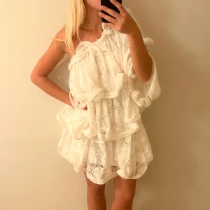 Vit spetsig oneshoulderklänning Positano - Superfin vit kortklänning i spets med volanger och oneshoulder-design. Klänningen har en romantisk och luftig känsla med mycket struktur och detaljer. Det är Lovisa Wallin collektion med NA-KD. Helt slut på hemsidan! (Endast andvänd en gång)