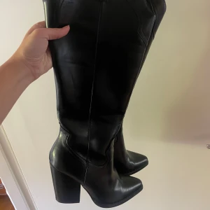 Svarta höga cowboy boots med klack - Säljer ett par svarta höga cowboy boots i skinnimitation med bred klack och spetsig tå. Snygga sömmar och klassisk westernstil, perfekta att styla med jeans eller klänning. Dragflik upptill för enkel påtagning.